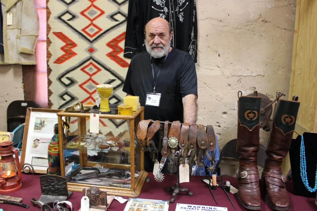 Bald vendor in black shirt standing at his booth with a glass display case of spurs and silver items, tooled leather belts, and a pair of tall star-badge cowboy boots with turquoise necklace at the 2018 CCG show