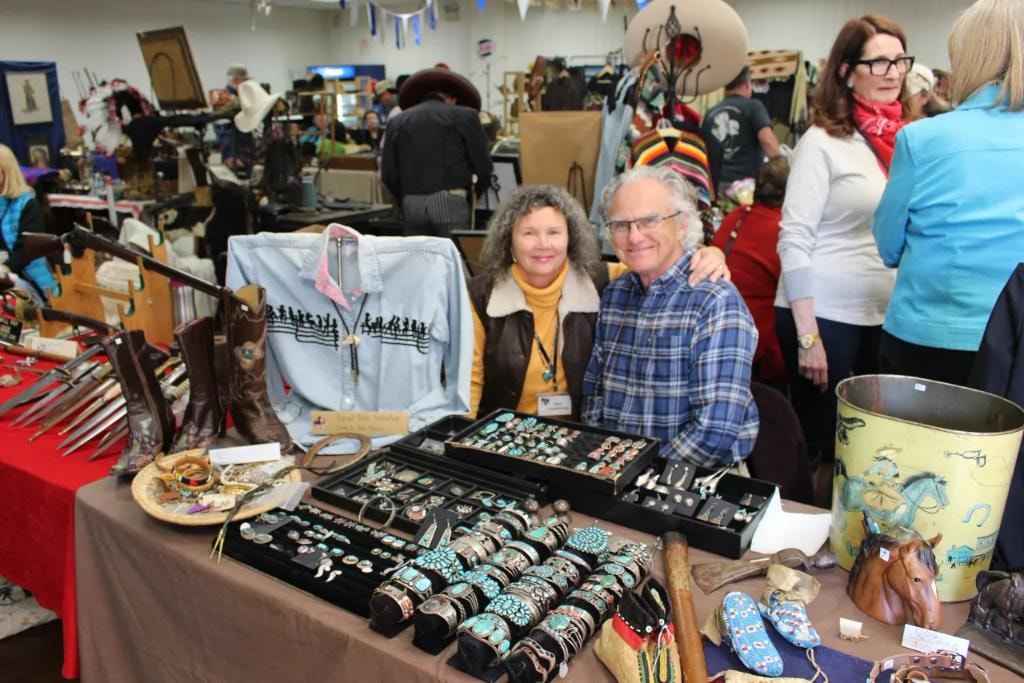 Smiling couple at their vendor booth displaying trays of turquoise and silver rings, cuff bracelets, and squash blossom necklaces alongside cowboy boots and a vintage western shirt at the 2018 CCG show