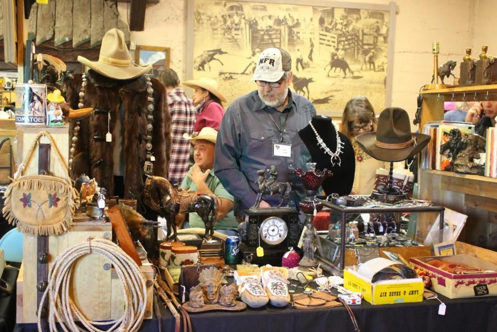 Man in NFR cap standing at his packed vendor booth with a bronze mantel clock, beaded moccasins, turquoise squash blossom necklace, and western collectibles at the 2018 CCG Prescott show