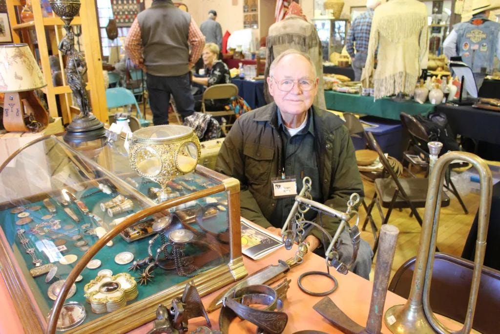 Older man seated at his vendor booth with a glass display case of silver spurs, coins, and buckles alongside antique horse bits, a brass bugle, and western collectibles at the 2018 CCG Prescott show