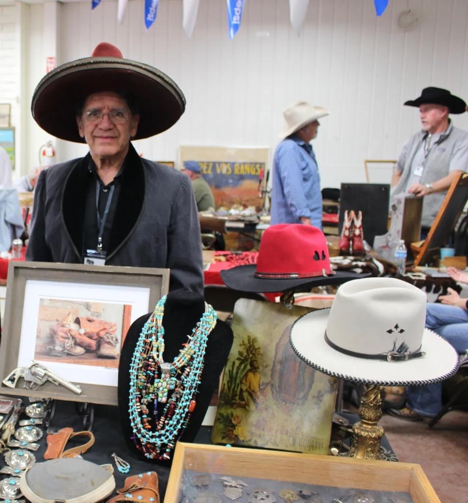 Man in a wide-brim sombrero at his vendor booth displaying red, black, and cream cowboy hats on stands alongside multi-strand turquoise necklaces, belt buckles, and western paintings at the 2018 CCG show