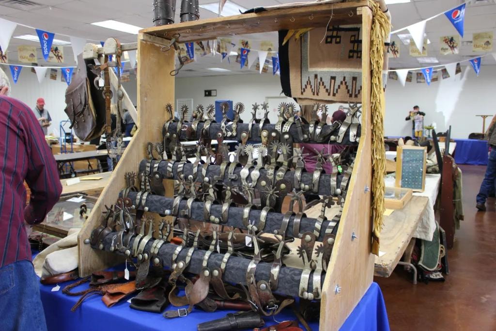 Large wooden spur rack packed with dozens of antique western spurs on leather straps displayed under Pepsi banners and CCG pennants at the 2018 CCG Prescott Rodeo Grounds show