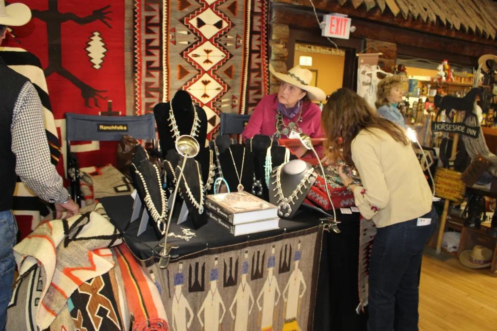 Woman in white cowboy hat behind a table of squash blossom necklaces on black velvet busts with large Navajo rugs hanging on the wall and a Horseshoe Inn sign in the background at the 2018 CCG Prescott show