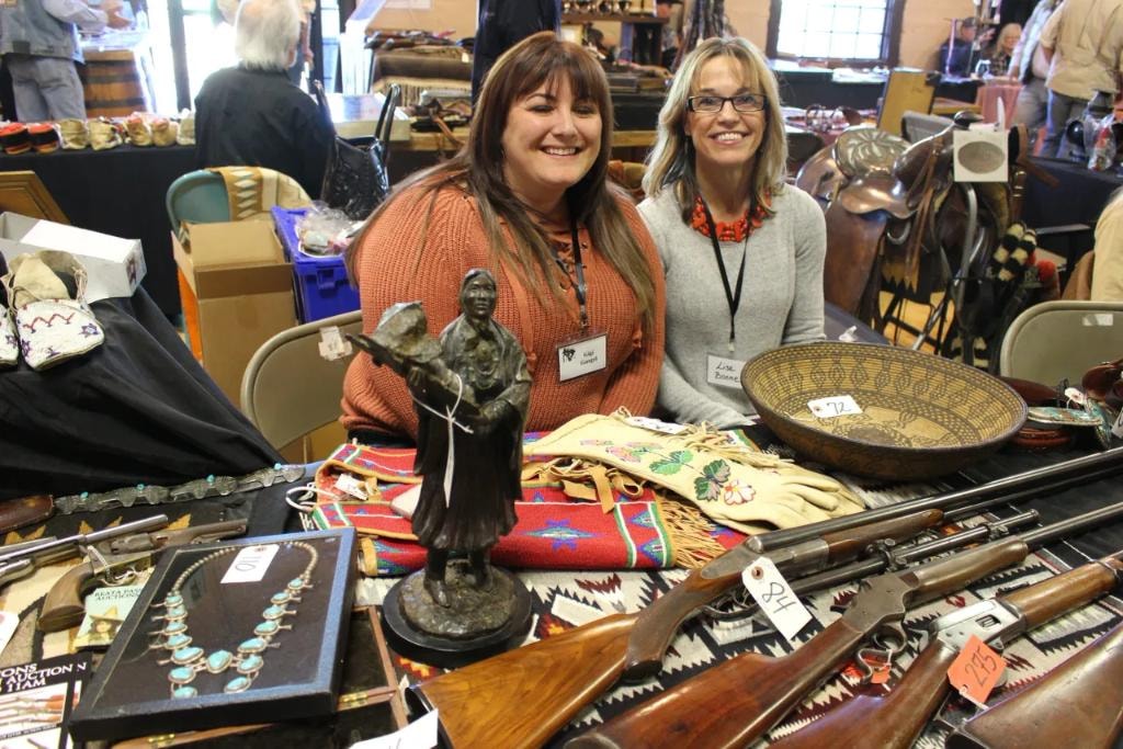Two smiling women vendors at their booth with a bronze Native American woman sculpture, turquoise squash blossom necklace, antique rifles, beaded gloves, and a woven Navajo blanket at the 2018 CCG show