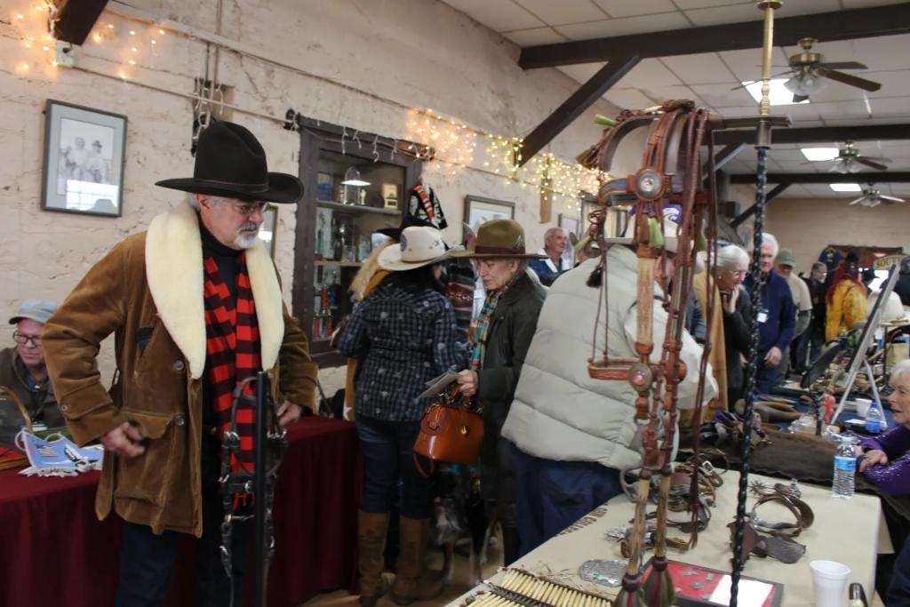 Man in black cowboy hat and shearling coat browsing a vendor table of antique horse tack, spurs, and bridles with a crowd of collectors behind him at the 2019 CCG Prescott Rodeo Grounds show