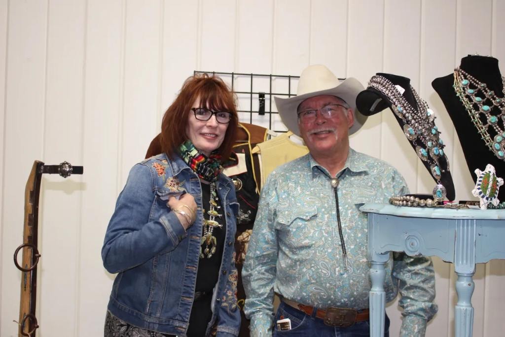 Woman in denim jacket and man in paisley western shirt and white cowboy hat standing at their vendor booth displaying squash blossom necklaces and turquoise jewelry