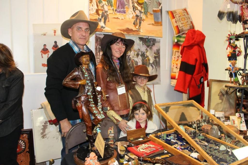 Family of four — man, woman, boy, and girl in cowboy hats — at a vendor booth filled with western paintings, kachina dolls, Navajo textiles, and a bronze cowboy sculpture