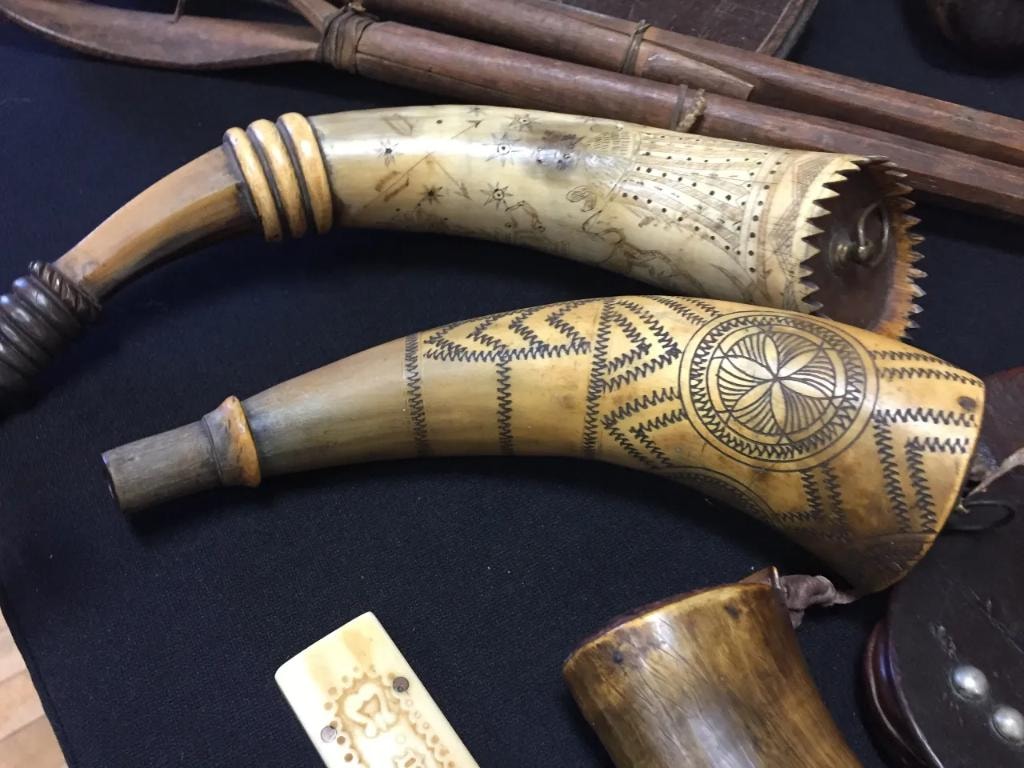 Two antique scrimshaw powder horns with intricate engraved geometric and celestial patterns displayed on a black cloth table alongside carved bone artifacts