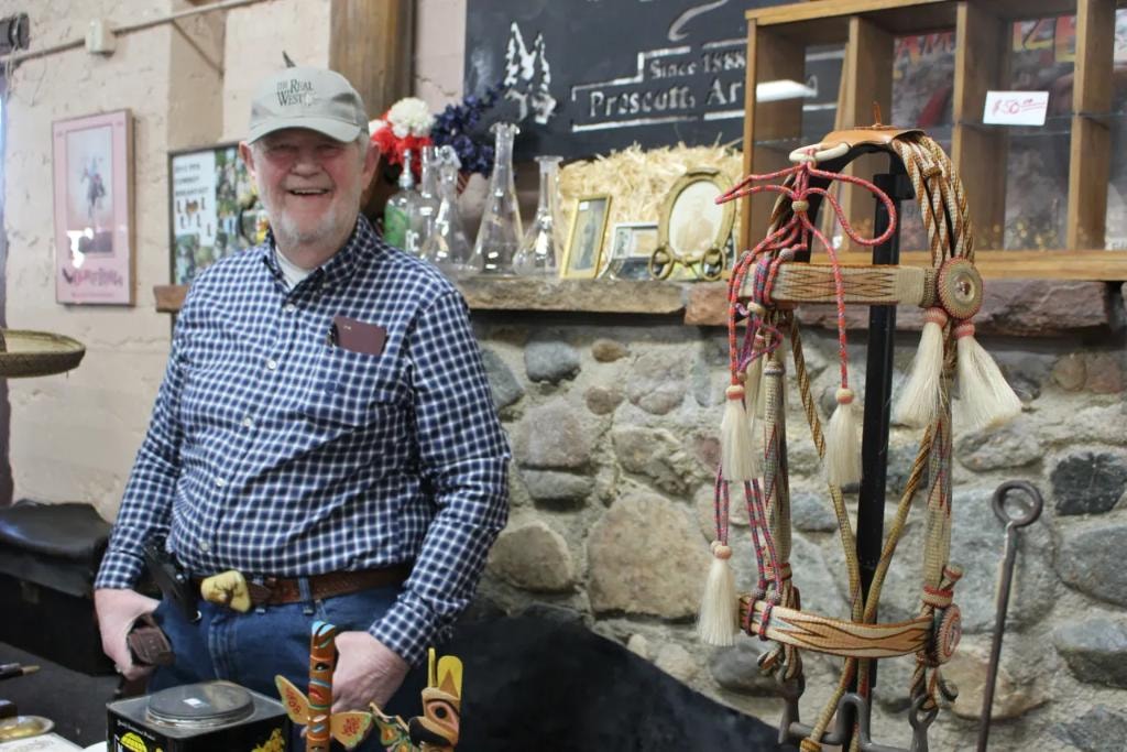 Smiling vendor in a plaid shirt and Real West cap seated at his booth beside a display of colorful braided horsehair reins and tassels against a stone wall at the 2019 CCG Prescott show