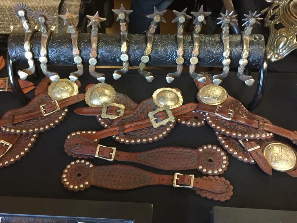 Row of antique western spurs mounted on a tooled leather roll with studded spur straps and large engraved brass conchos displayed on a black table