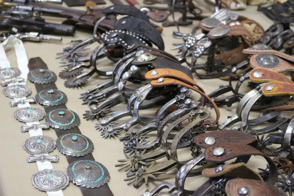 Table covered in antique western spurs, bits, and silver concho belts with turquoise stones alongside vintage firearms at the 2019 CCG show