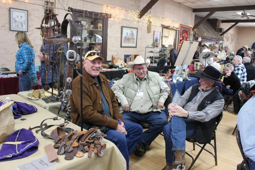Three men relaxing and chatting at a vendor booth with antique spurs and leather straps on the table and a packed show floor of collectors visible behind them at the 2019 CCG Prescott Rodeo Grounds