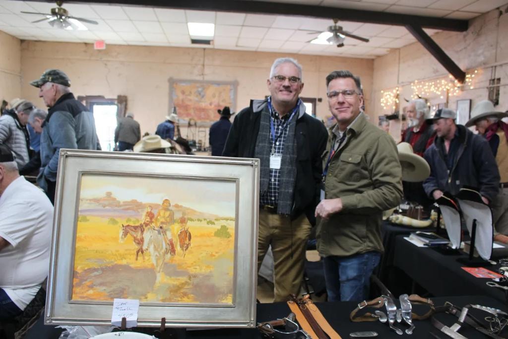 Two men smiling at a vendor booth with a large framed western oil painting of Native Americans on horseback in a golden landscape at the 2019 CCG Prescott show