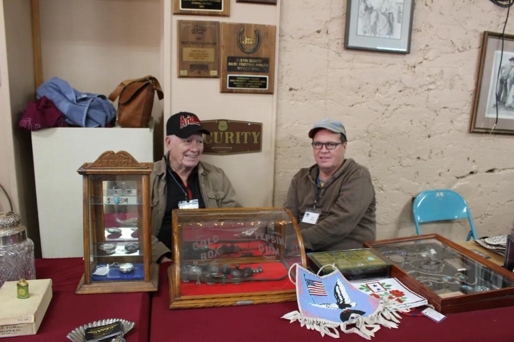Two men seated at a vendor booth with glass display cases containing pocket watches, belt buckles, and a vintage Gold Box Pepsin Gum tin at the 2019 CCG show