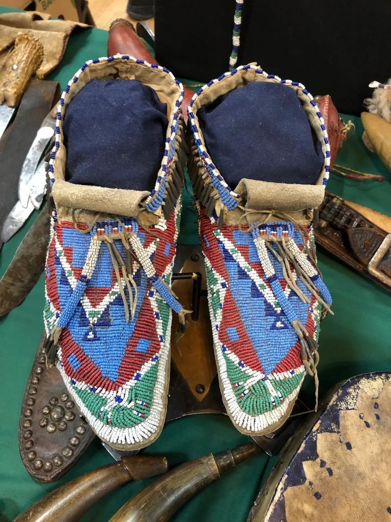 Pair of Native American beaded moccasins with blue, red, white, and green geometric diamond patterns on a green cloth table surrounded by western collectibles