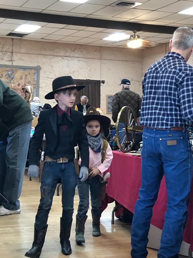 Two children dressed in full western cowboy costumes — older boy in black hat and duster, younger girl in pink shirt and bandana — walking the show floor at the 2020 CCG Prescott Rodeo Grounds event