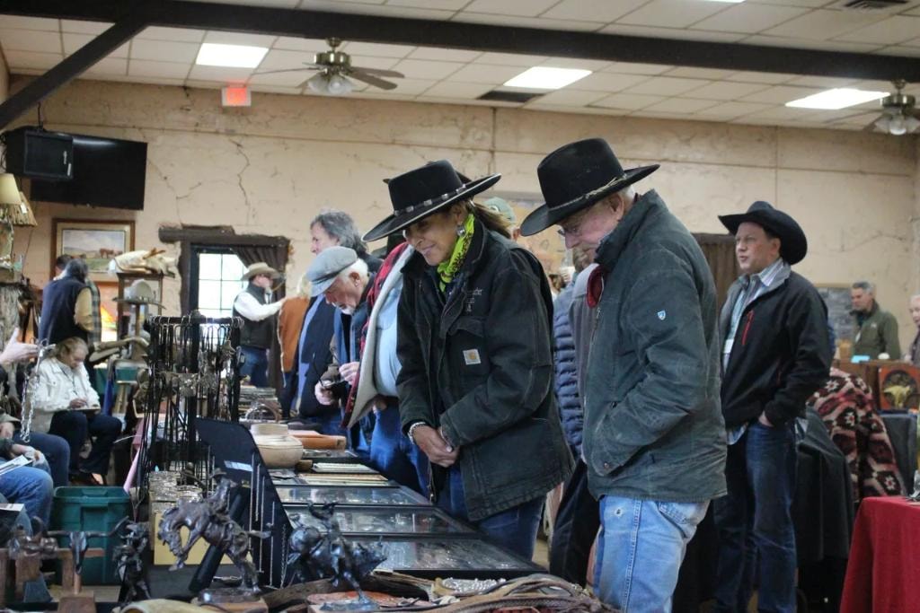 Two collectors in black cowboy hats leaning over a table of bronze sculptures and western antiques at the 2020 CCG show at Prescott Rodeo Grounds