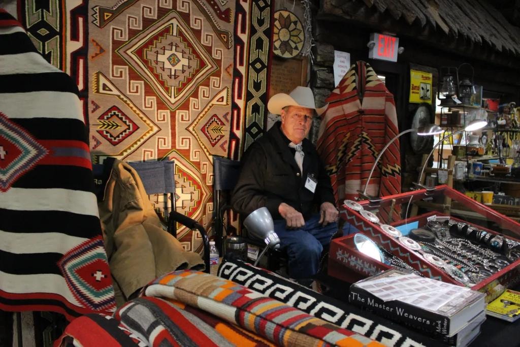 Cowboy vendor in white hat seated at his booth surrounded by large Navajo rugs, Pendleton blankets, and a glass case of silver jewelry