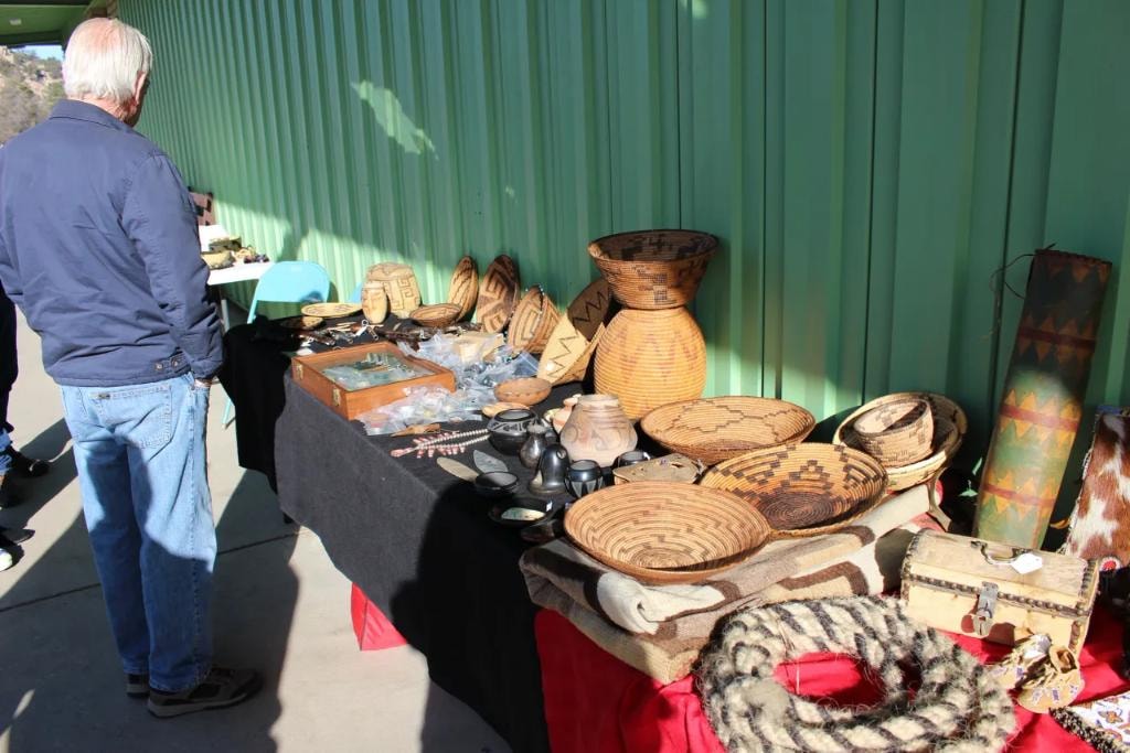 Outdoor vendor table at Prescott Rodeo Grounds covered in Native American woven baskets, pottery, and collectibles against a green metal building