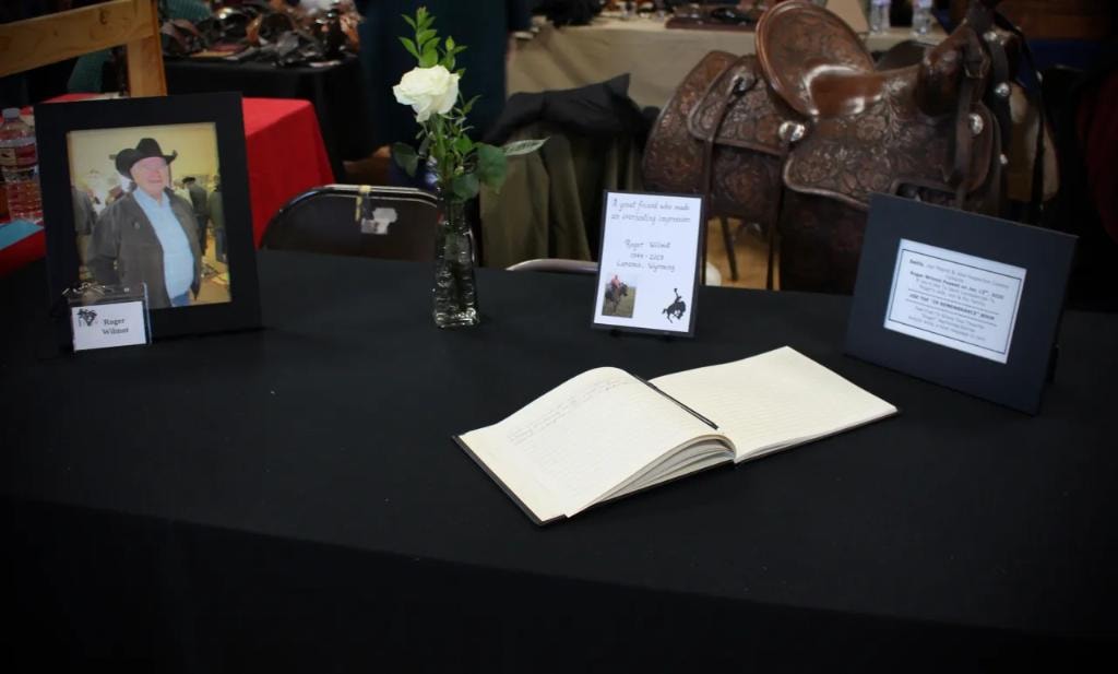 Memorial table for Roger Wilmot (1944–2019) of Laramie Wyoming with framed portrait, white rose, remembrance book, and a western saddle in the background