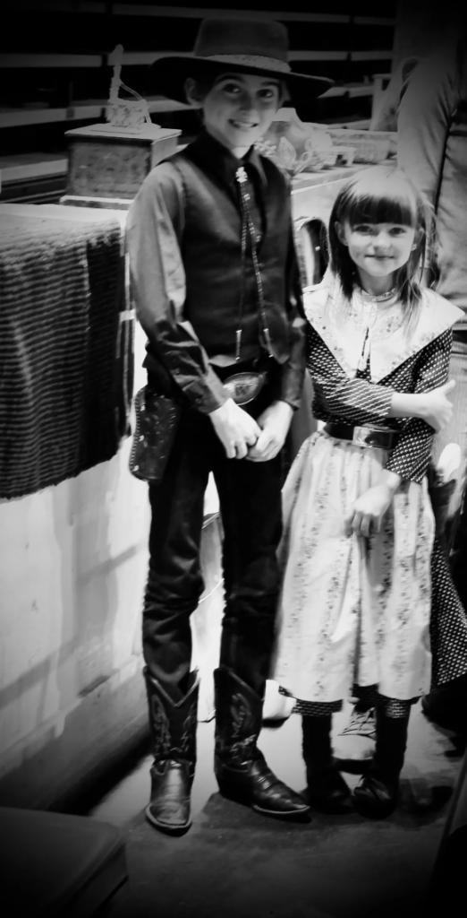Black and white photo of two children dressed in vintage western costumes — boy in cowboy vest and girl in pioneer dress