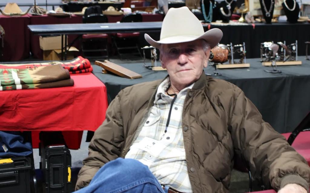 Older man in white cowboy hat seated at his vendor booth surrounded by Pendleton blankets and western collectibles