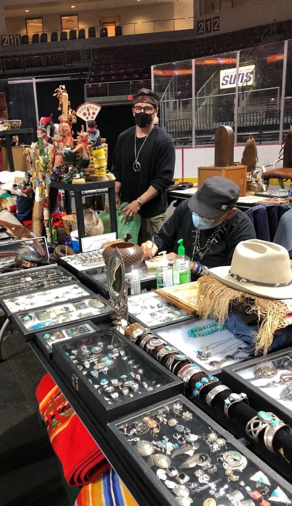 Two Native American vendors in masks behind glass cases filled with turquoise and silver jewelry, kachina dolls, and collectibles at the 2021 CCG show