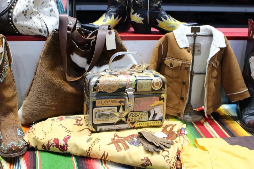 Vintage Yellowstone National Park tin lunchbox surrounded by cowhide bag, cowboy boots, sherpa jacket, and serape blanket at a vendor booth