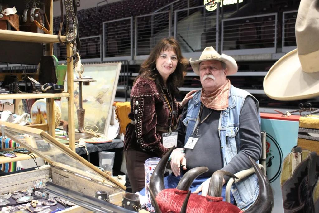 Woman in studded velvet jacket and man in cowboy hat with bandana posing together at their western collectibles booth