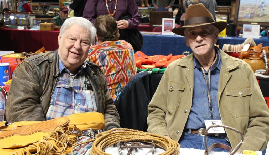 Two older men seated side by side at a vendor booth with tooled leather bags, rope, and colorful textiles