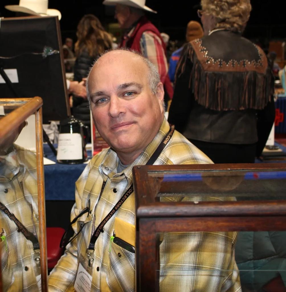 Man in plaid shirt seated at his vendor booth with a wooden display case, fringed leather vest visible behind him