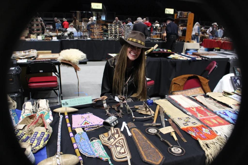 Smiling woman vendor in cowboy hat behind a table of Native American collectibles and beadwork