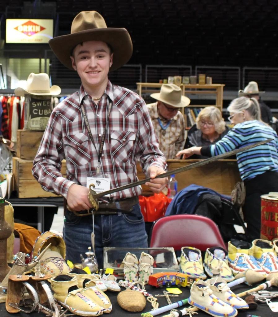 Smiling young man in cowboy hat holding an antique sword at a booth covered in beaded moccasins and Native American items