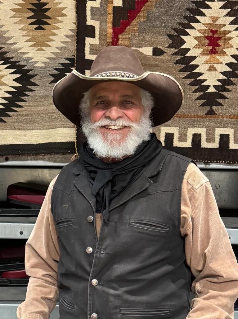 Smiling cowboy in brown hat and leather vest in front of Navajo rugs